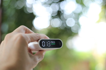 measuring outdoors air quality. A hand holding particulate matter 2.5 (pm.2.5) sensor. harmful small dust detector indicated healthy acceptable air quality. blur background of blue green plants.
