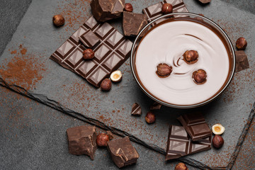 glass bowl of chocolate cream or melted chocolate, pieces of chocolate and hazelnuts on dark concrete background