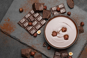glass bowl of chocolate cream or melted chocolate, pieces of chocolate and hazelnuts on dark concrete background
