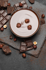 glass bowl of chocolate cream or melted chocolate, pieces of chocolate and hazelnuts on dark concrete background