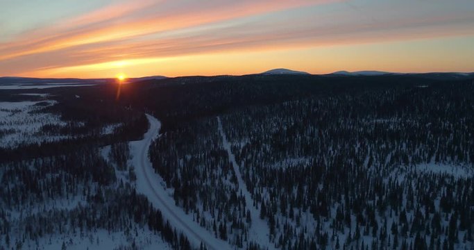 drone shot, following a vehicle, on a frozen route, between finnish woods and polar wilderness, in Pallas-yllastunturi national park, at the arctic circle, at sunset, on a sunny, winter evening dus