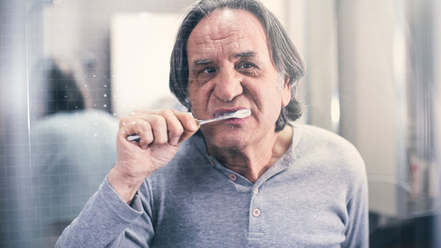 Old Man Brushing Teeth In Front Of The Mirror