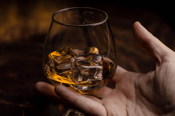 Whiskey / cognac glass with ice in a hand on a wooden background. Dark backdrop.