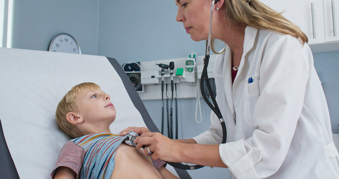 Female pediatrician using stethoscope to check the heartbeat of little boy on exam table. Woman doctor and sick child patient in clinic performing regular check up
