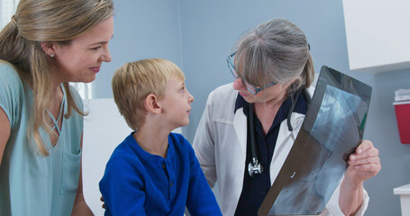 Senior woman pediatrician showing x-ray of spine to child patient and his mother. Little boy at doctors office for scoliosis