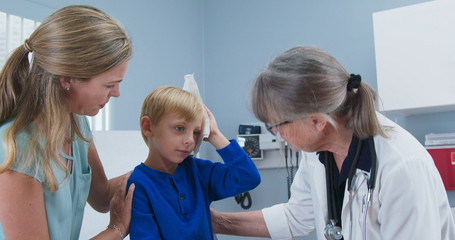 Child at pediatricians office holding ice pack on his head with mother and doctor in room. Little...