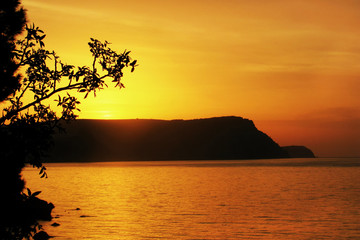 Fototapeta na wymiar Orange sunset over sea. Sun sets beautifully over ridge of mountain. Seascape with a silhouette of a hill in Bay.