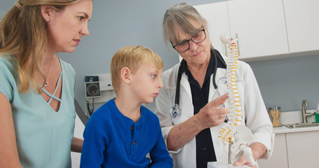 Senior woman pediatrician with spine model talking to little boy patient and his mother about...