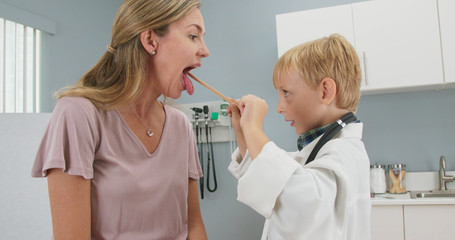 Cute little boy pretending to be a doctor with his mother while in pediatricians office. Child wearing oversized lab coat and stethoscope using tongue depressor