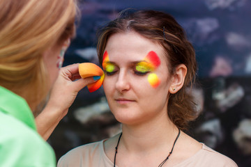 Girl put a picture on her face. Make-up of paints on a body for a carnival. An adult woman is...