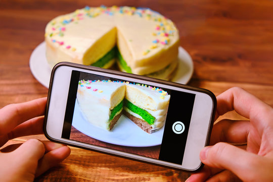 Women's hands with the phone taking pictures of the cake. Blogger shoots food for instagram. Create a photo for posting on the Internet. White phone and a tri-color cake on a wooden table.