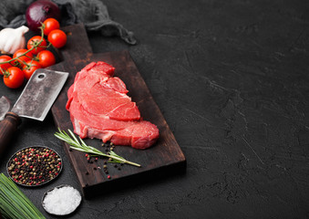 Fresh raw organic slice of braising steak fillet on chopping board with meat hatchets on black stone background. Red onion, tomatoes with salt and pepper. Space for text