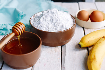 Ingridients on the wooden white table. Banana, flour, egg, honey. Bread. Raw.