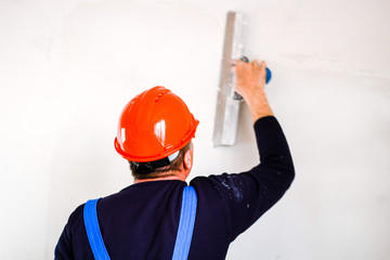 A builder in an orange construction cup in his hands with a spatula and a white putty. Blue form...