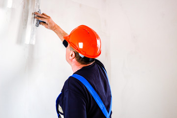 A builder in an orange construction cup in his hands with a spatula and a white putty. Blue form...