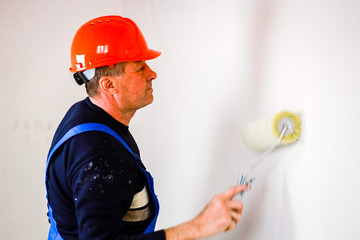 A builder in an orange construction cup in his hands with a spatula and a white putty. Blue form...