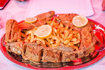 Delicious fried trout fish and catfish pieces, fried potatoes, decorated with lemon slices on disposable paper plate. Fresh fish from Tisza river