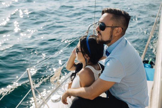 A girl and a guy of Asian appearance are sitting on the deck of the yacht, embracing each other. The couple looks to the horizon.