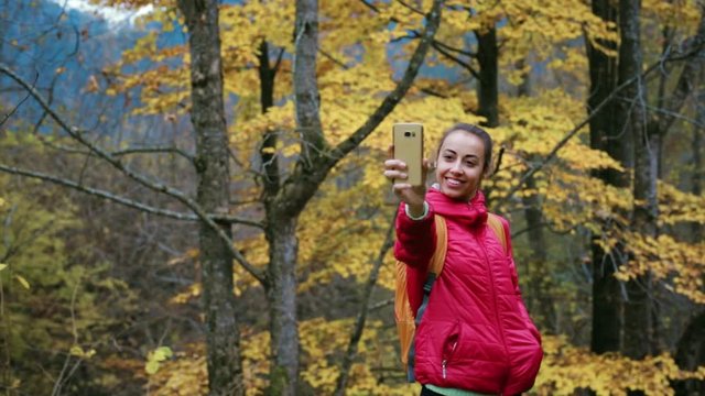 pretty woman hiker in red jacket with small backpack is standing on the yellow autumn woods background using phone and making selfie.