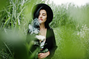 Sensual smoker girl all in black, red lips and hat. Goth dramatic woman hold white chrysanthemum...