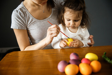 Fototapeta premium A young woman and her daughter are painting on an easter egg together. The girl is holding the egg while a woman is painting on it. Isolated on gray background.
