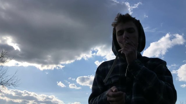 handsome young guy in hood smoking cigarette outdoors on the blue cloudy sky background. Slow motion