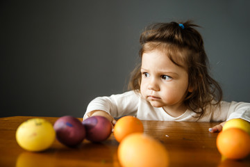 Fototapeta na wymiar Angry baby girl is sitting behind the table with a bunch of colored eggs laying on it. She is looking away. Isolated on gray background.