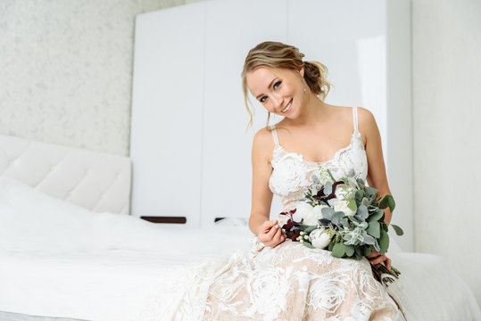 Happy bride with a cute smile and with a wedding bouquet in her hand poses in a room with a white interior