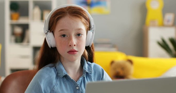 Portrait of the cute Caucasian red haired teen girl in the headphones sitting in front of the laptop computer screen in the nice room, then smiling to the camera. Close up.