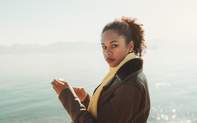Portrait of a young curly-hair Brazilian tourist female standing near the river in warm clothes on a sunny day and using her smartphone to tell friends in a chat about her springtime travel
