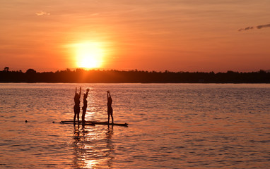 Silhouette of three girls doing yoga trainer balancing on the water on the paddle board over beautiful orange sunset background, Gili Trawangan Island, Indonesia