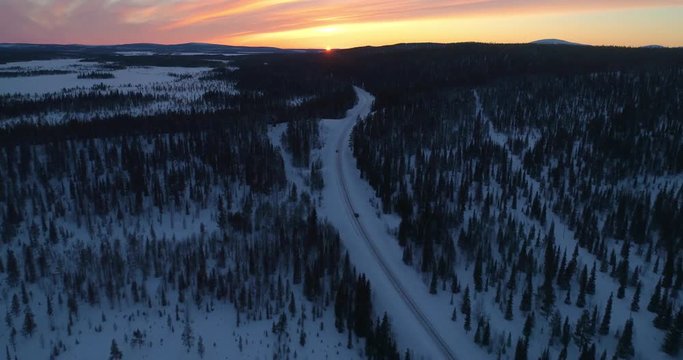 drone shot, following a vehicle, on a frozen route, between finnish woods and polar wilderness, in Pallas-yllastunturi national park, at the arctic circle, at sunset, on a sunny, winter evening dus