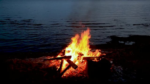 Home style footage. Sound. Twenty nine 29 seconds of bonfire or campfire for Guy Fawkes theme by a body of water at dusk. Increase of flames with leaves added.