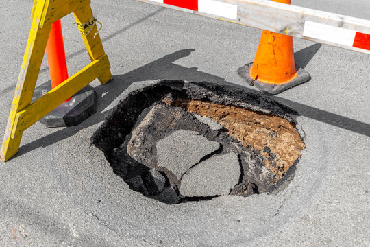 A large, deep pothole in a road. Part of a traffic cone and sawhorse visible blocking the hole.