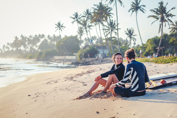 Young beautiful loving couple friends are sitting on the sand by the ocean with surfboards and...