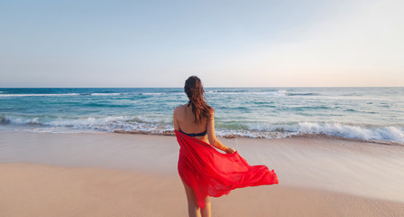 Fototapeta na wymiar Beautiful young brunette woman with long hair in a bikini with a bright red scarf in her hands on the ocean at sunset