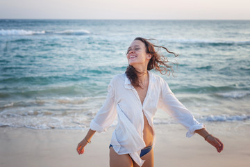 Beautiful happy young brunette woman with long hair in a white shirt enjoys enjoying life near the ocean at sunset
