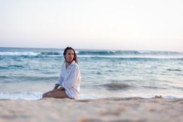 Fototapeta na wymiar Beautiful young brunette woman with long hair in a white shirt sitting on the ocean at sunset