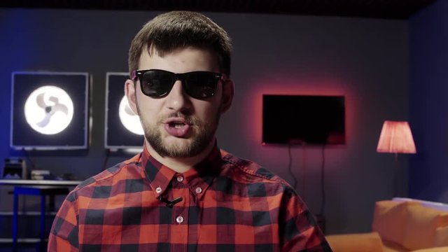 Interesing cute young bearded guy wearing stylish sunglasses and red checkered shirt with small wireless mic looks at camera and emotionally speaks in modern furnished room.