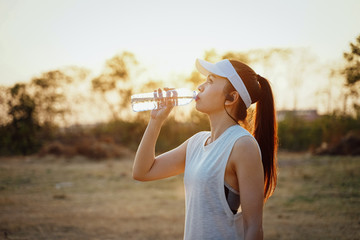 Young woman runner wears white sport vest, white cap, black short pants drinks mineral water after running.