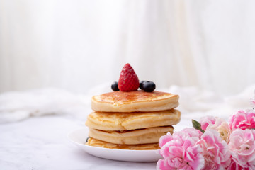 Pancakes on white plate topped with strawberry, rasberry and black currants and honey syrup and pink blooming flower in blackground.