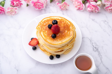 Pancakes on white plate topped with strawberry, rasberry and black currants and honey syrup and pink blooming flower in blackground.