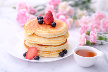 Pancakes on white plate topped with strawberry, rasberry and black currants and honey syrup and pink blooming flower in blackground.