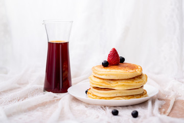 Front view of Pancakes on white plate topped with strawberry, rasberry and black currants and honey syrup and pink blooming flower in blackground.