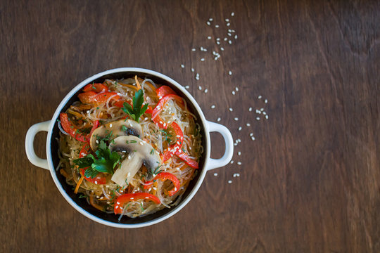 glass noodles with vegetables and mushrooms in a plate on a brown background. Top view, close-up, copy space