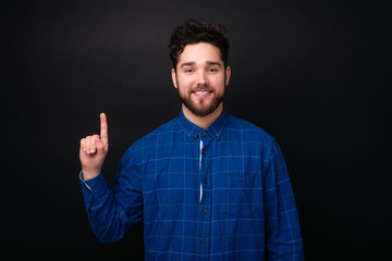 Happy smiling bearded young man looking at camer and pointing up with finger over dark background