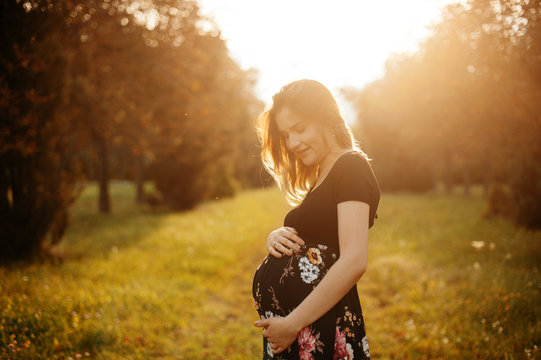 Beautiful tender mood photo of pregnancy. Young pregnant woman, holds hands on belly, in park on sunset