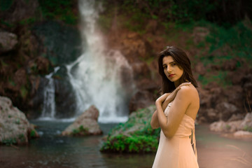 Fototapeta premium Sexy woman posing on the rock among green tropical plants and beside beautiful waterfall with blue water. Fairy tale.