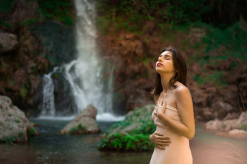 Sexy woman posing on the rock among green tropical plants and beside beautiful waterfall with blue water. Fairy tale.