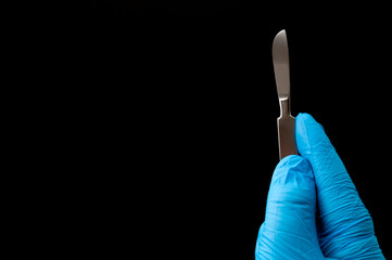 Surgical instruments in operating room and doctor performing surgery concept theme with a surgeon hand wearing blue latex gloves and holding a scalpel isolated on black background with copy space
